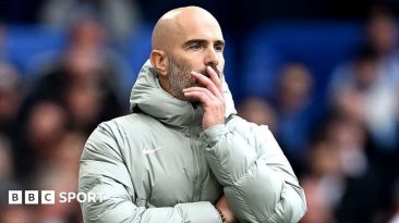 Enzo Maresca, looks on during the Premier League match between Chelsea and Brighton at Stamford Bridge. He is wearing a grey coat and holding his face.