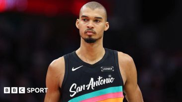 Victor Wembanyama wearing a vest that is black with blue, pink and orange stripes and the words San Antonio across the front