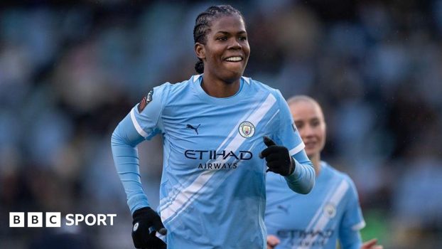 Bunny Shaw, wearing a sky blue Man City shirt and black gloves, grins as she runs