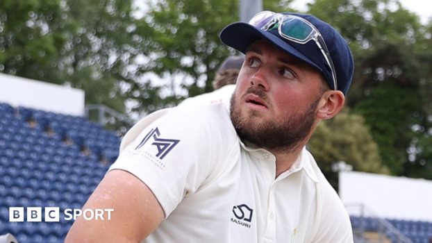Tom Norton seated on the boundary with glasses on top of cap looks over his shoulder towards the stands at Sophia Gardens