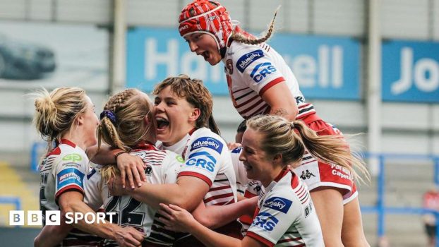 Wigan Warriors women players, in white kit with red trim, surround try-scorer Ruby Hunter, with one jumping on the shoulders of a team-mate